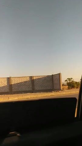The video shows a view from inside a moving vehicle, possibly a car, looking out the window. The scenery consists of a roadside with sparse vegetation, a low dirt embankment, and electrical utility poles. A motorcycle carrying two people briefly passes by. The sky is clear and light blue. The overall impression is of a dry, arid landscape.
