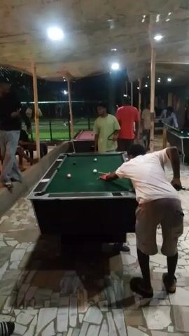The video shows several people playing pool at an outdoor bar or recreation area. In the foreground, a person in a white t-shirt and shorts is playing pool on a green table, taking a shot with a cue stick. Behind him, two other people stand by another pool table covered in red cloth. Several other individuals sit in chairs in the background, either watching the game or relaxing. The area is covered by a partially translucent roof supported by posts, and outdoor lighting is visible. The floor is tiled with geometric patterns. The scene appears to be at night or dusk.