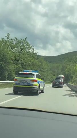 The video shows a white police SUV with blue emergency lights on its roof, driving on a two-lane asphalt road. Its rear license plate reads "PA 9131 MA" and "POLICE" is visible below the plate. The police vehicle is followed by a dark-colored SUV with a roof rack, and further ahead, a large truck.

The road is surrounded by dense green trees and hills, suggesting a rural or mountainous setting. The sky is overcast with gray clouds. The video is filmed from inside another vehicle, looking forward through the windshield. The vehicles are moving in the same direction.