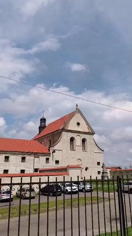 The video shows a cloudy sky above a large, old building with a red tiled roof. The building appears to be a church or a monastery, with a tall bell tower visible. In front of the building, there is a black metal fence, and behind it, several cars are parked. The scene is static, with no movement or action taking place. The lighting suggests it is daytime.