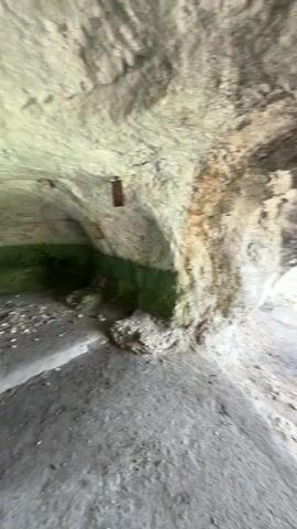 The camera is inside a rock shelter or cave, panning from left to right. The interior has rough, light-colored rock walls and ceiling, with some darker patches and a green area on the left wall. The floor is flat and appears to be concrete or smoothed rock. As the camera pans right, it reveals a large opening looking out onto a bright green landscape with numerous trees and bushes under a clear sky. A dark-colored SUV is visible on a dirt road that winds through the foliage. A colorful piece of fabric, possibly a hammock, hangs near the right side of the cave opening.