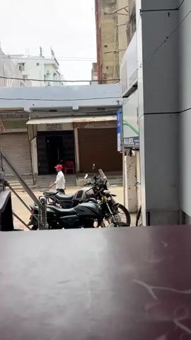 This video shows a street scene in front of shops, with a person walking in the background. Two motorcycles are parked in the foreground, and a railing is visible on the left. The shops appear to be closed, with their shutters down. The street is paved, and there are buildings on either side. The sky is overcast.