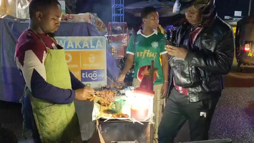 Here is a description of the video.

The video shows a street scene at night. A man in an apron is cooking meat skewers on a grill at a small outdoor food stall. Another man, wearing a motorcycle helmet and a black jacket, is looking at his phone. A third man in a green and white soccer jersey stands nearby. In the background, there is a line of auto rickshaws, and a bus drives by. A sign with the word "WAKALA" is visible on the food stall. The scene appears to be a busy roadside area with vehicles and people passing by.