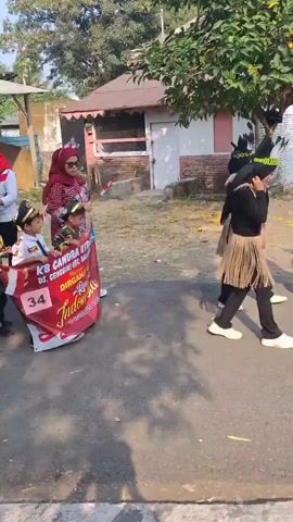 A group of young children dressed in various costumes, including military uniforms and dresses, are participating in a parade. They are accompanied by adults, some of whom are wearing red and white attire and headscarves, holding Indonesian flags. A banner with Indonesian text and the number "34" is displayed. The scene appears to be outdoors, possibly in a village or school setting, with buildings and trees visible in the background. The children and adults are walking along a paved road. The atmosphere seems festive and celebratory.