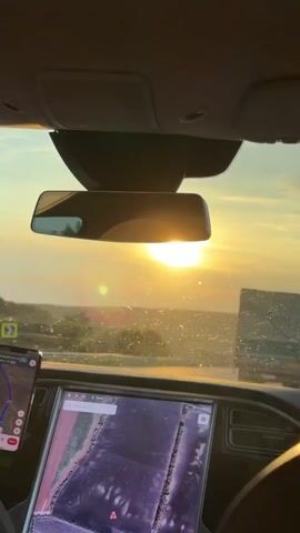 The video is a first-person view from inside a vehicle, looking through the windshield and at the rearview mirror. The sun is visible in the distance, low on the horizon, casting a bright glare and creating lens flares. The sky is a warm yellow and orange, indicating either sunrise or sunset. The interior of the car is partially visible, showing the dashboard with a large screen displaying a map application. A smartphone is mounted on the left side of the dashboard, also showing a navigation app. Outside the car, the landscape appears somewhat hazy and distant, with hints of trees or other vegetation. There are also other vehicles visible in the distance on what appears to be a road. The windshield has some dirt or debris on it, which is illuminated by the sun. The overall impression is of a scenic drive during golden hour.