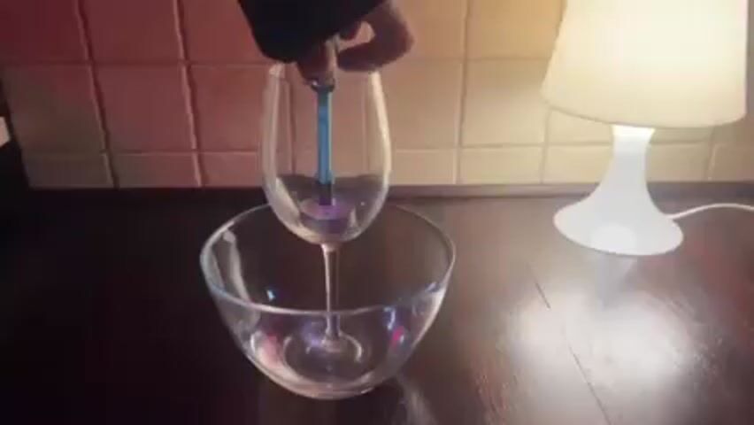 Here's a description of the video:
A person's hand removes a blue candle snuffer from atop a wine glass containing a lit candle, which sits inside a glass bowl. The bowl rests on a dark wooden table, illuminated by a white lamp on the right. A tiled wall forms the backdrop.