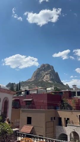 The video shows a daytime shot of a mountainous landscape. In the foreground is a rooftop patio with a decorative wooden bench and potted plants. Below, various buildings with red and beige rooftops are visible. In the background, a large, gray mountain dominates the scene. The sky is bright blue with scattered white clouds. The overall atmosphere is sunny and peaceful.