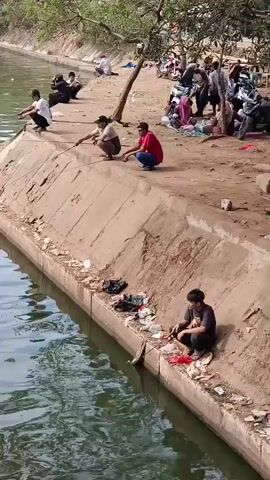 The video shows an outdoor scene with a body of water on the left and a dirt bank on the right. Several people are fishing from the bank. In the foreground, a person is crouched down and holding a fishing rod. Further up the bank, a group of people are also fishing, with some sitting and others crouching. In the background, there are trees, motorcycles, and more people gathered. The bank is littered with trash. The water appears to be murky green.