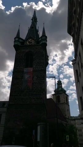 The video shows a low-angle, static shot of two church towers against a cloudy sky. The left tower is larger and darker, made of stone, with a clock face visible near the top. A banner with an image of a fish and some text hangs from the tower. A second, smaller tower with a green dome is visible behind and to the right of the first tower. The right side of the frame shows a portion of a modern building. The lighting suggests it is daytime, but the clouds are casting shadows. There are some indistinct shapes at the bottom of the frame which may be people or vehicles, but they are not clearly discernible. The overall impression is of an urban environment with historical architecture.