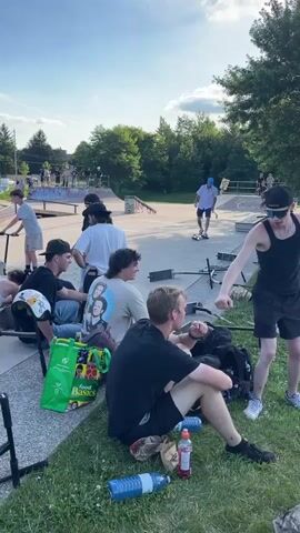 The video depicts a group of young people gathered at an outdoor skate park or recreational area. The scene appears to be set in the late afternoon or early evening, with the sun casting long shadows. Several individuals are seated on the grass, some holding drinks or food. Others are standing, engaged in conversation or observing activities. One person is seen riding a scooter, and there are several scooters and skateboards scattered around the area. In the background, there is a skate park structure with ramps and other features, as well as a grassy field with trees and a soccer goal in the distance. The overall atmosphere seems relaxed and social, with the group appearing to be enjoying a casual gathering.