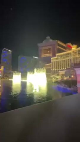 At night, illuminated fountains spray water high in the air in front of buildings, including a hotel and a casino in Las Vegas. The water is lit up and shines brightly, reflecting in the water beneath. One building in the background is tall with a red top, and another is lit up with blue lights. The water is black with white reflections, and the sky is dark. The camera is still, and there is a grey wall in the foreground.