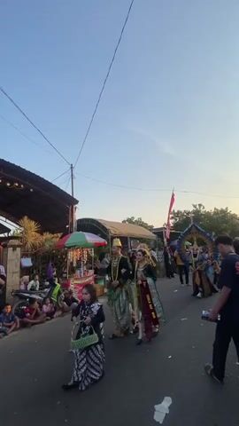 The video shows a street scene with people participating in a parade or procession. In the foreground, a young girl in traditional clothing walks towards the camera, carrying a basket. Behind her, a couple dressed in elaborate traditional attire, likely a bride and groom, walks together, arm in arm. They are surrounded by other participants and onlookers. The environment appears to be a village or small town, with buildings and temporary stalls lining the street. The sky is clear and blue, suggesting it is daytime. The overall atmosphere seems festive and celebratory.