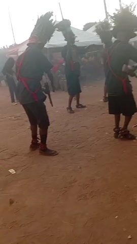 The video shows a group of people performing a traditional dance or ritual in what appears to be an outdoor setting. The participants, who seem to be predominantly male, are wearing dark shorts, shirts, and distinctive headwear, possibly made of feathers or similar natural materials. Red bands or sashes are worn across their chests, and some are carrying what appear to be rifles or long wooden staffs. They are moving around on a reddish-brown dirt surface. In the background, there are tents, people watching, and utility poles. A person in the foreground is holding a smartphone up, possibly recording or taking photos of the event.