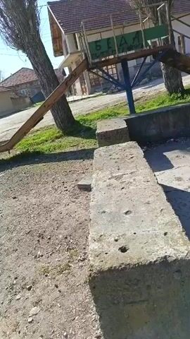 The video shows a view of a small village on a bright sunny day. The camera pans across a dirt and gravel ground, with patches of green grass growing sparsely. A concrete structure sits in the foreground, possibly a low wall or bench. To the left, an old, rusty metal slide stands beside a tall, bare tree. In the background, there are several modest buildings with tiled roofs, typical of a rural setting. A sign with illegible writing is visible atop a structure supported by blue metal poles. The sky is clear and blue. The scene is quiet and appears deserted.