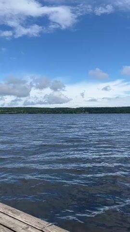This is a low-angle, first-person perspective video taken from a wooden dock on a body of water. The water is dark blue and slightly choppy with visible ripples. In the distance, a treeline forms the horizon, and beyond that, a few scattered houses are visible. The sky is a bright blue with scattered white clouds, some of which are beginning to gather and become more grey. The camera pans slightly to the right, revealing more of the lake and sky. The dock itself is made of weathered wooden planks, and two vertical wooden posts are visible in the foreground on the right side of the frame. The overall scene is tranquil and evokes a sense of being outdoors on a summer day.