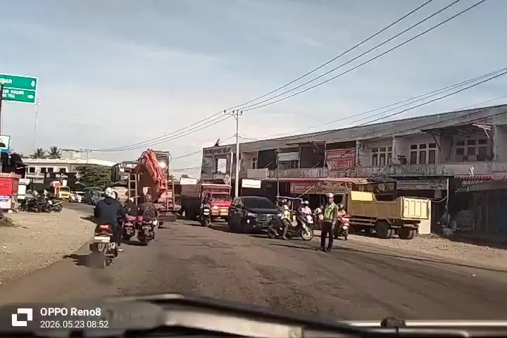The video shows a busy street scene in what appears to be a developing area. There are several vehicles including cars, trucks, and motorcycles on the road. A large piece of construction equipment, possibly an excavator, is being transported on a trailer pulled by a truck. A traffic officer is seen managing the flow of traffic. Buildings and shops line the side of the road. The sky is partly cloudy. The video was shot with an OPPO Reno 8 at 08:52 on May 23, 2026.