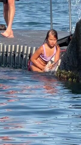 A young girl in a purple swimsuit is sitting on the edge of a floating dock, partially submerged in blue water. She is holding a stick and appears to be playing in the water. Her hair is wet and she has a focused expression on her face. In the background, other people are visible on the dock, including a man whose legs are in the frame and two boys. The sun is shining brightly, indicating it is likely daytime, and the water is calm with ripples. The dock is made of modular plastic sections and is tied to a large, rocky structure covered in green moss or algae on the right side of the frame. The overall scene suggests a leisurely day at a waterfront location, possibly a lake or the sea, with children enjoying recreational activities.