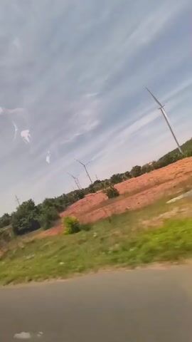 The video shows a view from a moving vehicle, looking out at a landscape with wind turbines. The landscape is characterized by rolling hills, some covered in reddish-brown earth and others with green vegetation. Several wind turbines, with their tall white towers and blades, are visible in the distance. The sky is blue with wispy white clouds. The road in the foreground is asphalt. There are no people or animals clearly visible. The overall impression is of a rural or semi-rural area with renewable energy infrastructure.