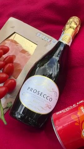 A picnic scene unfolds on a red blanket spread across a grassy lawn. In the foreground, on the blanket, are several food and drink items: a bottle of Prosecco with a gold foil top and a white label, a pack of smoked salmon, a cluster of cherry tomatoes, and a red can of Fanta. The camera pans out to reveal a larger picnic setting in the background, where a group of people are seated at a picnic table under a large tree. The environment is a park or a field with lush green grass and trees under a bright, partly cloudy sky.