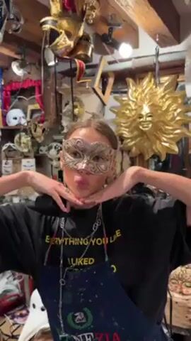 A young woman wearing a black t-shirt, a denim apron with the word "VENEZIA" written on it, and a gold and silver mask, poses for a video in a shop filled with masks. The woman has her hands raised to her chin and is making a kissing face. Various masks are displayed in the background, including a gold sun mask. The shop is cluttered with masks and other items hanging from the ceiling and walls.