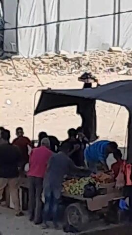 A group of people are gathered around a cart under a black tarp. The cart is laden with various items, including what appears to be produce and textiles. Some individuals are interacting with the items on the cart, while others stand nearby, observing or engaged in conversation. The environment appears to be outdoors, with a sandy ground and a large, white fabric structure in the background, possibly a tent or a temporary shelter. The lighting suggests daytime.