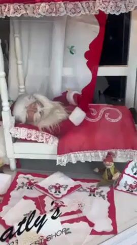The video shows a shop window display decorated with Christmas themes. A mannequin dressed as Santa Claus is lying in a white bed with red bedding adorned with lace and embroidered with an "S" design. A red curtain drapes next to the bed. On the floor of the display, there are embroidered placemats and napkins featuring Santa Claus designs. A small elf figurine sits near the bed. The text "Latly's Gift Shop 2023" is superimposed on the lower portion of the display. The lighting is bright, suggesting the scene is taken during the daytime.