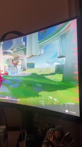 The video shows a person playing a video game on a computer monitor. The game appears to be a colorful, futuristic, third-person shooter or action game. The game's environment is bright and airy, with large white structures and lush green fields. There are various visual effects in the game, including glowing particles and bright explosions of light, suggesting combat is taking place. In the foreground of the game, there are character models with vibrant colors.

The player's health is displayed as 493/493. There is also text in Russian on the screen, including "НАЗАД" (back) and "ИЗ 5 НОКАУТОВ!" (out of 5 knockouts!). A list of player names is visible in the upper right corner, some of which include "Veidman," "KingKobold," and "Cracked Choom."

The monitor is a curved, widescreen display. Below the monitor, partially visible, are various objects including a can, a keyboard, and what appears to be a collectible figurine of a warrior-like character. The lighting in the room suggests it is nighttime or the room is dimly lit, with the computer monitor being the primary light source. The video is taken from a slightly elevated angle, looking down at the monitor. The setting is likely a home environment. The overall mood conveyed by the game's visuals is energetic and action-oriented.