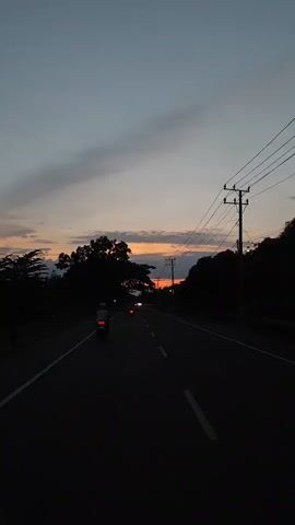 The video shows a straight road at dusk. The sky is a gradient of dark grey to orange, with the orange glow concentrated on the horizon, indicating a setting sun. There are trees silhouetted on either side of the road, with an electrical pole and wires on the right. Several vehicles, including one or two motorcycles, can be seen driving away into the distance. The road is paved, with white lane markings. The overall scene is dark.