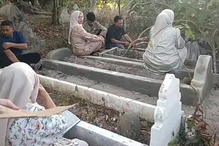 The video shows a group of six individuals gathered in what appears to be a cemetery. Three women are wearing hijabs, and they are seated on or near the grave markers. One young woman, with a black hijab, stands at the front left holding a piece of cardboard to block the sun. A second woman, wearing a light-colored hijab and floral top, sits nearby, reading from a book with Arabic script. The remaining four adults are sitting amongst the graves. Two of them are sitting cross-legged. One individual appears to be tending to the grave. The environment suggests an outdoor setting with trees.