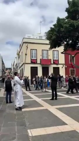 The video shows a crowded public square in front of colorful buildings. The square is paved with light-colored stones forming a grid pattern. A man in a white religious robe stands in the foreground, holding a tall cross and facing away from the camera, toward a crowd of people. The man appears to be conducting a religious ceremony or leading a procession. Most of the crowd is standing. There is a man in a suit in the center of the frame, but otherwise, there is no particular focus on any individual in the crowd. The weather is partly cloudy, and there is a large tree in the background.