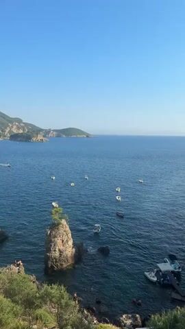 The video shows a body of water with several boats. In the foreground, there is a rocky island with a tree on top. There are also people visible on the shore and in the water. In the background, there are hills and a clear blue sky. The video appears to be filmed from an elevated position, looking out over the water.