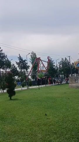 A swing ride in an amusement park swings back and forth. The ride is red and yellow and is set against a backdrop of trees and a cloudy sky. The camera is positioned on a green lawn looking towards the ride. The ride is the central focus of the video, with the camera panning slightly to follow its motion.