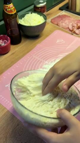 A pair of hands is shown preparing dough in a glass bowl. Flour is sprinkled onto a pink silicone mat beside the bowl. The hands are seen lifting the dough from the bowl, which appears to be sticky and elastic. In the background, there are various ingredients for pizza, including a bowl of shredded cheese, a jar of sauce, and slices of salami. The scene takes place on a wooden countertop.