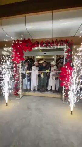 The video captures a festive ribbon-cutting ceremony taking place at the entrance of a building, likely in Kandahar, Afghanistan. Approximately 8 to 10 men, mostly dressed in traditional white shalwar kameez with some wearing turbans or skullcaps, are gathered in front of the entrance.

The entrance is framed by a white archway elaborately decorated with abundant bright red artificial roses, creating a celebratory atmosphere. A yellow or gold ribbon is stretched across the doorway, held by several of the men. In the center, one man holds a pair of scissors, poised to cut the ribbon.

Adding to the grand occasion, two large sparkler fountains are actively spraying bright white and silver sparks upwards on either side of the archway. The scene appears to be taking place in the evening or at night, given the dramatic effect of the sparklers and the reliance on artificial lighting from within the building. The interior visible through the entrance seems brightly lit. The ground in the foreground is a plain gray surface. No discernible text is visible on the building or decorations. The overall mood is celebratory and formal, marking the inauguration of what appears to be a new commercial or public establishment.