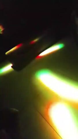 The video shows a dark interior space, possibly a club or event venue, with various colored lights flashing and moving around. Initially, a red light illuminates the lower portion of a dark object. The lights shift to blues, greens, and pinks, and the space appears more expansive with distant figures moving. The lighting then becomes predominantly blue. The sound of music is present throughout.
