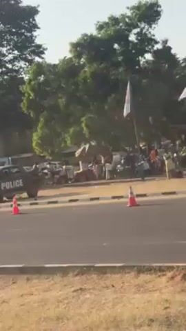 The video shows two separate outdoor scenes with cars and crowds. 

The first scene shows a silver car with damage to the front passenger side. There is a police vehicle in the background along with an orange traffic cone. A crowd of people is visible in the background behind tents and flags. The setting appears to be a grassy area next to a paved road. 

The second scene shows a road winding uphill past a group of people and cars. The background features a dry, hillside. A grey car is driving on the road toward the camera and a red car is driving away from the camera.