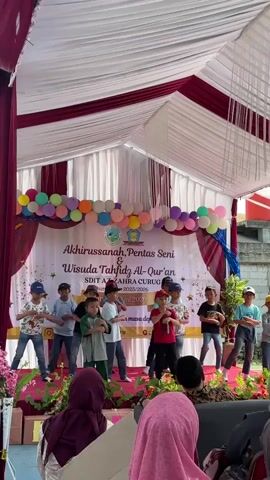 A group of young boys, dressed in casual clothing and some wearing baseball caps, are performing a coordinated dance routine on a stage set up outdoors. The stage is decorated with balloons and a banner that reads "AKHIRUSSANAH, PENTAS SENI & WISUDA TAHFIDZ AL-QUR'AN SDIT AZ-ZAHRA CURUG". The boys are positioned across the stage, some in front and others in the back, moving their arms and bodies in sync with the music. In the foreground, out of focus, are audience members seated, mostly women wearing headscarves. The setting appears to be a school event or celebration, possibly a graduation ceremony.