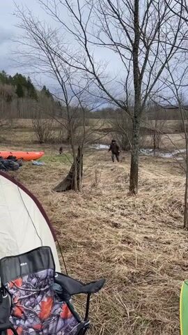 The video shows a campsite with a tent and a folding chair in the foreground. A man walks across a grassy, marshy field in the midground, carrying an object in his right hand. There is an orange inflatable kayak lying on the ground near some camping gear. Behind the field are trees and a cloudy sky. The scene appears to be a rural area on a cool, overcast day.