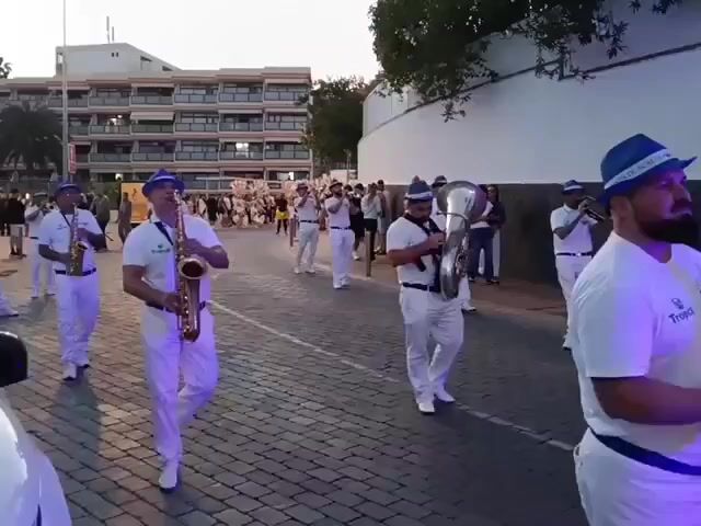 A marching band is performing on a brick-paved street. The band members, predominantly male, are dressed in white trousers, white shirts, and blue hats with white ribbons. They are playing various instruments, including saxophones, trumpets, and a tuba. Some band members have "BANDA DE ACAETE" and "Tropical" printed on their shirts. They are walking in formation, playing their instruments in sync. A few bystanders are observing the performance. In the background, there is a multi-story building with a hotel-like appearance and a white wall with a green tree behind it. The lighting suggests it might be evening, with a slightly dim sky. A white car is partially visible on the left side of the frame.