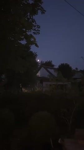 This is a vertical video clip of a rural scene at dusk or night. The moon is visible in the sky, appearing as a bright, round object. In the foreground, there are dark silhouettes of trees and bushes. In the background, a white-gabled house with a dark roof is visible, partially obscured by the trees. The sky is a deep blue, transitioning to black towards the end of the clip. The overall impression is one of quiet and darkness.