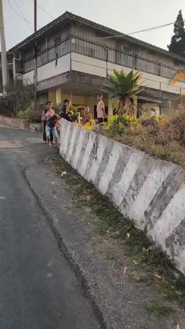 A group of people, including children, are gathered near a building with a concrete barrier in the foreground. The sun is shining, and there are trees and vegetation in the background. The scene appears to be outdoors in a rural or semi-rural setting.