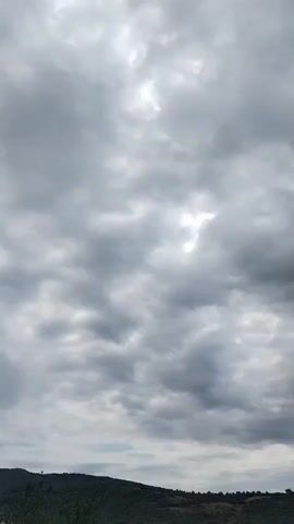 The video shows a landscape dominated by a cloudy sky. Below the sky, there is a dark, hilly terrain covered in vegetation. The lighting suggests it is daytime, but the clouds are dense, obscuring direct sunlight. There are no visible people or objects in motion within the frame. The scene is static and focuses on the atmospheric conditions over the landscape.