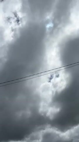 The video shows a vertical shot of a cloudy sky with two power lines running horizontally across the frame. The clouds are mostly dark and heavy, suggesting an overcast or stormy day. In the lower portion of the video, the roofline and upper part of a wooden building are visible. A chimney stack can be seen on the roof, and a metal antenna is mounted on a pole near the chimney. The building appears to be somewhat aged. There are no people visible in the video. The environment appears to be rural or semi-rural, given the presence of the building and antenna against the natural backdrop of the sky.