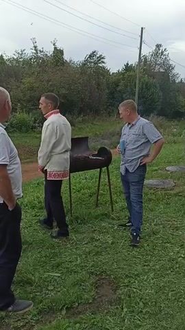 Three men are standing around a barbecue. One man in a white t-shirt and black pants stands with his back to the camera, his hands in his pockets. Another man, wearing a white shirt with red embroidery and dark pants, walks away from the barbecue. The third man, in a blue and white checkered shirt and blue jeans, stands near the barbecue holding a blue cup and looking at the other two men. The barbecue, a metal barrel on legs, is lit and has food cooking on it. A table with bottles and a pot is to the left of the barbecue, and a gray plastic bag is on the ground next to it. A silver car is parked behind the table. The scene is outdoors on a grassy area with trees and bushes in the background. The sky is overcast.