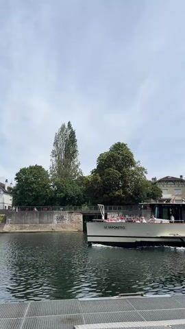 A boat named Le Vaporetto travels along a body of water. Several people are visible on the deck of the boat, some seated and some standing. The boat has a covered section with windows and a visible sign that reads "Garonne Boits Croisières". Trees and buildings are visible on the shore in the background, under a cloudy sky. The camera is positioned at water level, looking towards the boat as it moves away.