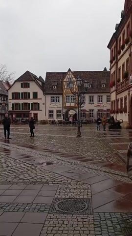 The video shows a rain-soaked town square in what appears to be Europe, possibly Germany, given the architecture. The ground is covered in cobblestones and large square tiles, reflecting the overcast sky. Several people are seen walking across the square, some carrying umbrellas. Buildings with traditional European architectural styles, including timber framing and steeply pitched roofs, surround the square. Some of the buildings have awnings and outdoor seating, suggesting cafes or restaurants. In the center of the square is a fountain. The camera moves slightly, revealing more of the square and the surrounding buildings. The overall atmosphere is quiet and subdued due to the rain and overcast weather.