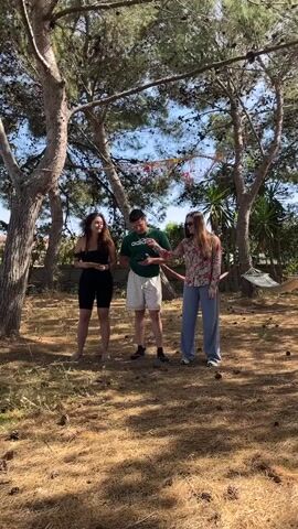 In a daytime shot, three people are standing outside on a plot of land beneath trees. A woman with long brown hair is on the left, dressed in black shorts and a black top and holding a yellow object in her hands. In the center is a man wearing a green t-shirt with the word ‘Adidas’ printed on it, and white shorts. To the right, a second woman, also with long brown hair, is wearing a light-colored floral blouse and loose-fitting light blue pants. Behind them, a hammock is visible, strung between two trees. The ground is covered with dry grass and pine cones. A string of decorations is hanging between the trees in the background. The scene is naturally lit, creating shadows under the trees and people.
