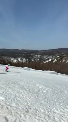 The video shows a snow-covered hillside on a sunny day with several people skiing. The skiers are wearing brightly colored outfits and are making their way down the hill. In the background, there is a landscape of trees and hills covered with snow. The sky is a clear blue.