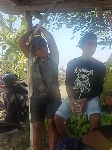 TWO MEN AND A BOY ARE SEEN UNDER A SHED WITH A THATCH ROOF. THE MAN ON THE LEFT IS WEARING A CAP AND A GRAY SHIRT. THE BOY IN THE CENTER IS WEARING A BLACK LONG-SLEEVED SHIRT AND BLUE SHORTS AND IS HOLDING A GLASS WITH A STRAW. THE MAN ON THE RIGHT IS WEARING A CAP AND A BLUE SHIRT AND IS HOLDING A CELL PHONE. THERE IS A MOTORCYCLE ON THE LEFT SIDE OF THE FRAME. THE BACKGROUND IS A RURAL SCENE WITH TREES AND VEGETATION.