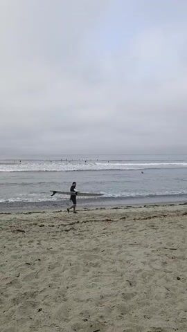 A person is running along a sandy beach carrying a surfboard. The ocean is to the left of the person, with waves breaking on the shore. The sky is overcast and cloudy. There are other people in the water further out, possibly also surfing. The sand is light brown and appears to be dry.
